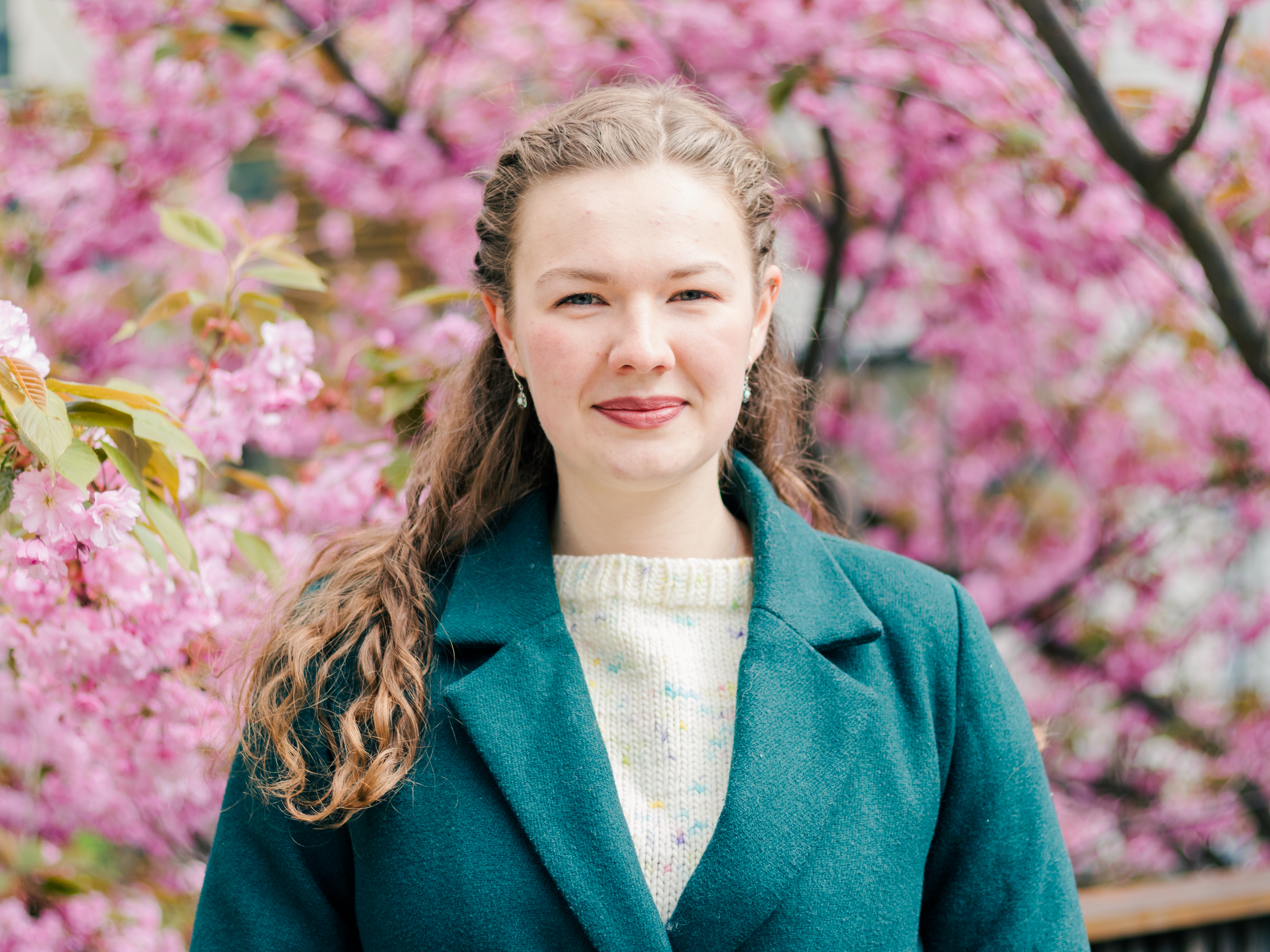 Headshot of PhD Fellow with green jacket and hand-knit sweater in front of pink blossoming tree